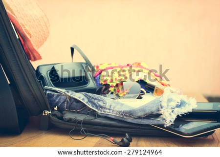 Open the suitcase with tourist things: women\'s hat, swimsuit, camera, denim shorts, dresses, sunglasses, perfumes, nail polish, mobile phone, tablet on wooden background.  Toned image.