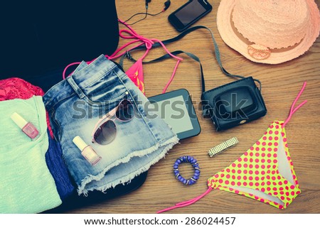 Open the suitcase with tourist things: women\'s hat, swimsuit, camera, denim shorts, dresses, sunglasses, perfumes, nail polish, mobile phone, tablet on wooden background. Toned image.