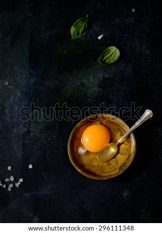 raw egg in a bowl, spoon, basil on a black background