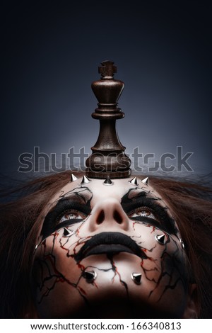 A creative photo of a queen with a painted pierced face and a chess king in her mouth.