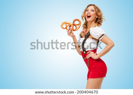 Young sexy Swiss woman wearing red jumper shorts with suspenders in a form of a traditional dirndl, eating two pretzels in her hand on blue background.