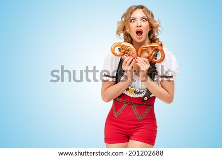 Young sexy Swiss woman wearing red jumper shorts with suspenders in a form of a traditional dirndl, holding with hunger two pretzels on blue background.