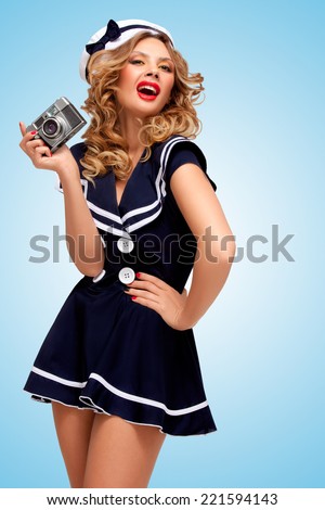 Retro photo of a glamorous pin-up sailor girl with an old vintage photo camera showing emotions on blue background.