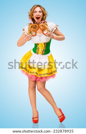 Young hungry sexy Oktoberfest woman wearing a traditional Bavarian dress dirndl holding two pretzels, and screaming on blue background.