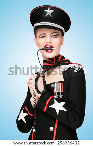 Sexy girl, dressed in a military uniform dress like a dominatrix, biting vintage unplugged music headphones on blue background.