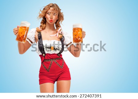Young surprised sexy Swiss woman wearing red jumper shorts with suspenders in a form of a traditional dirndl, holding two beer mugs on blue background.