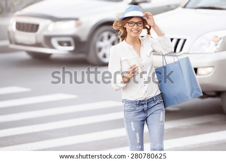 Happy young female student crossing the street with a coffee-to-go cup and tipping hat against urban city background.