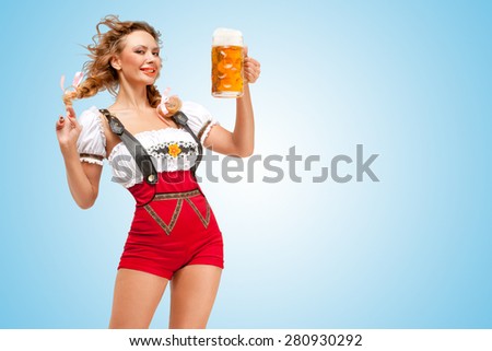 Young flirting sexy Swiss woman wearing red jumper shorts with suspenders in a form of a traditional dirndl, holding a beer mug on blue background.