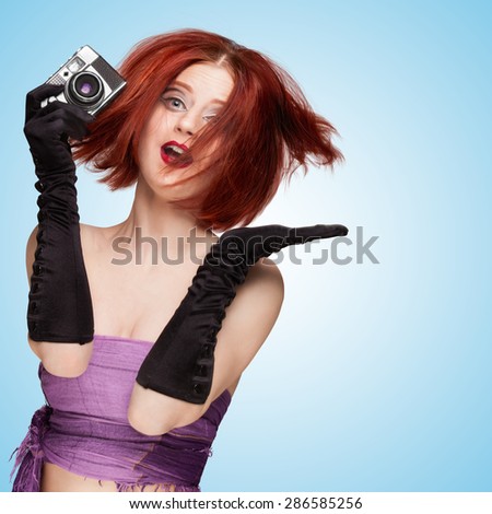 Glamorous emotional girl, wearing long gloves and disheveled hairstyle, holding an old vintage photo camera and jumping on blue background.