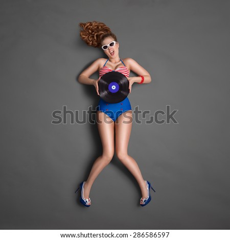 Beautiful pinup girl in retro bikini and sunglasses, holding an LP microgroove vinyl record and showing emotions on chalkboard background.
