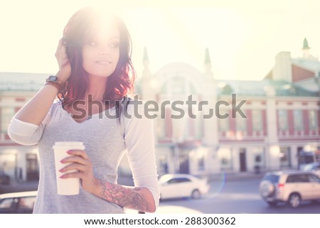 Beautiful young smiling woman with tattoo, holding a disposable takeaway cup and looking aside against urban city background.