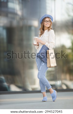 Happy young trendy woman drinking take away coffee, standing on the stairs with shopping bags, and looking aside with smile against urban city background.