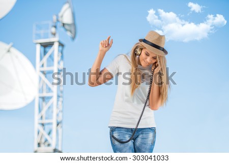 Happy young woman in hat listening to the music in vintage music headphones and dancing against background of satellite dish that receives wireless signals from satellites.