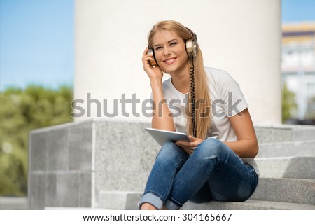 Happy young woman listening to the music in vintage music headphones, surfing internet on a tablet pc and sitting on stairs against urban city background.