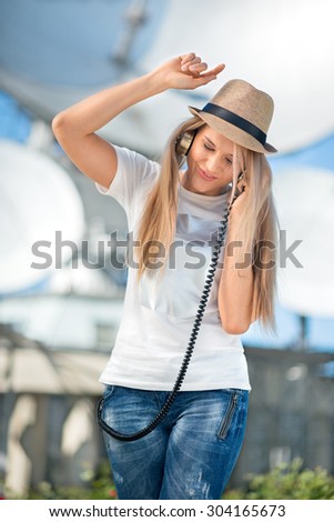 Happy young woman in hat listening to the music in vintage music headphones and dancing against background of satellite dishes that receive wireless signals from satellites.
