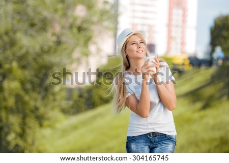 Beautiful young woman walking with a disposable coffee cup, drinking coffee, and smiling against urban nature background.