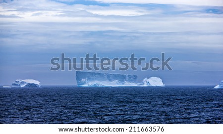 Antarctic iceberg floating in the ocean. Blue gradient between water, clouds, sky and snow.