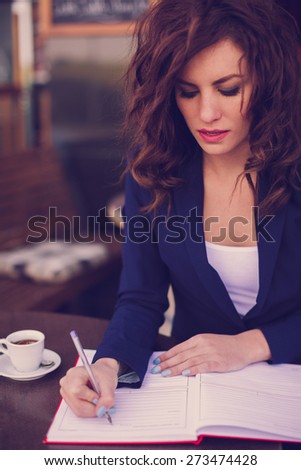 Young business woman sitting at the table and writing in a notebook. On table she has cup of coffee