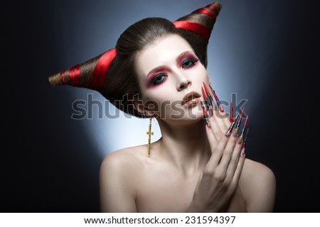 The girl in an image of the demon-tempter with long nails and haircut in the form of horns. Picture taken in the studio on a gray background.