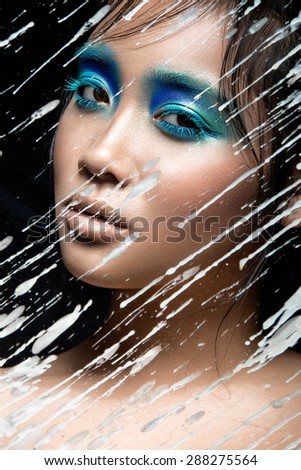 Beautiful Asian girl with bright blue make-up behind glass with drops of wax. Beauty face. Picture taken in the studio on a black background.