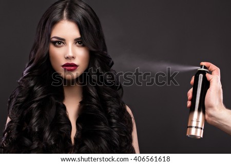 Beautiful brunette model with curls, classic makeup and red lips with a bottle of hair products. The beauty of the face. Portrait shot in the studio on a gray background.
