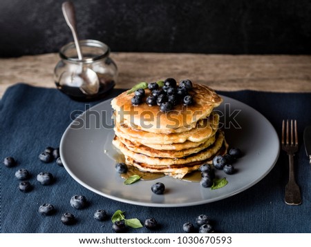 image shows a homemade fluffy pancake with blueberry on the top; situation is decorated with rustiv wooden table, placemat, silver cutlery, honey glass