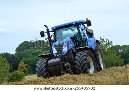 A stationary blue tractor is parked on the edge of a field of barley against an overcast sky.