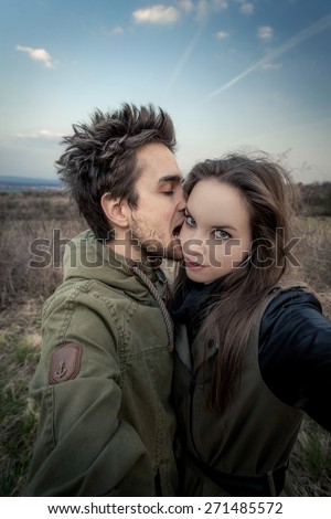 Man kissing / biting woman\'s face. Couple stretching hands to the camera which bought Selfie. In the background withered vegetation with blue sky. Photo in vintage style.
