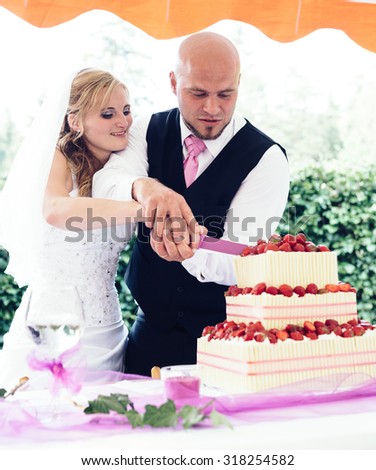 Bride and groom, hand in hand, are slicing the wedding cake with strawberries.(Focused on eyes of newlyweds, shallow depth of field)