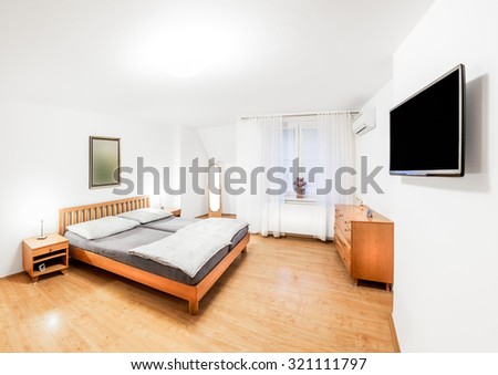 Ordinary bedroom of the house in wooden style with double bed, bedside tables, mirror, chest of drawers and a modern TV. Bedroom is lit by bright light mounted on the ceiling and two bedside lamps.