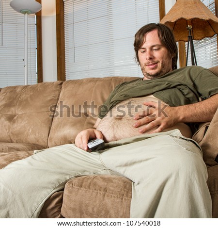 A large belly and a man smiling at it while being lazy with his TV remote in hand. Shows a couch potato or a man with a sedentary lifestyle .