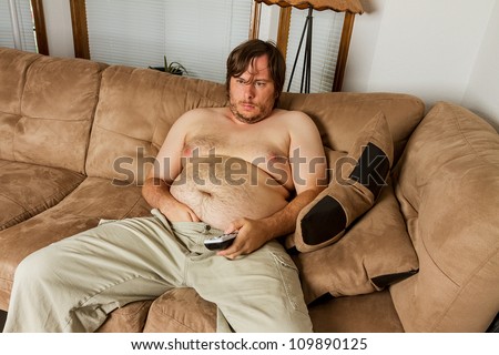 Fat obese man  on the couch with remote in one hand and the other hand shoved down the front of his pants. Shows obesity due to lack of exercise.