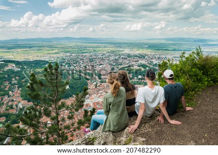 A group of four traveling friends sitting on the edge of a cliff and watching city from above. Brasov City, Romania viewed from Mount Tampa. Tourism concept for travel agency stock image