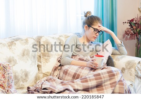 Image of a teenage girl feeling sick, sitting covered on the couch and drinking tea. Depressed young woman showing early Ebola symptoms.