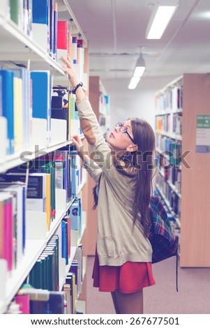 Attractive young female student searching for books in the library, Lifestyle image of girl reaching for a book, with a vintage, blue instagram filter applied. Knowledge is power concept.