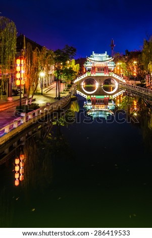 Chinese bridge over the canal with crystal clear reflection in the water. Beautiful lights and colorful decorations in Chinese village. China lit up at night, popular travel destination.