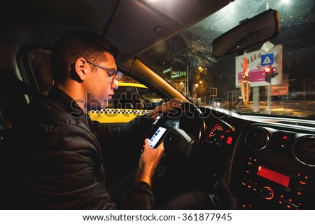 Young man texting and driving. Teenage driver on his cellphone while driving his car at night. Typing on the smartphone at the traffic lights. Dangerous driving concept.