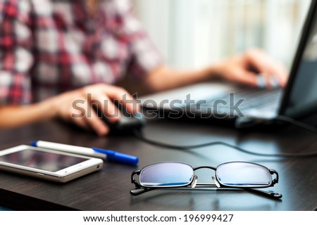 Glasses on the table with cell phone and student at work on the laptop