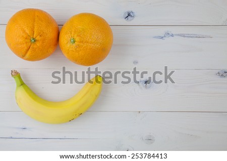 Two oranges and a banana that resembles two eyes and a smile lying on a white wooden table.