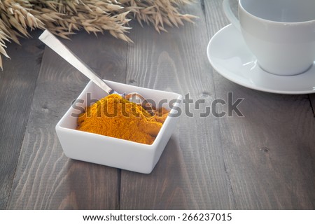 Turmeric in a white bowl with a spoon. The bowl stands on a dark gray wooden table where it is a white cup and grain lies in the background.