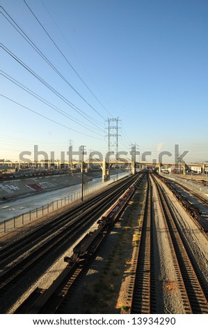Railroad tracks and power lines converge next to the Los Angeles River in the early morning.  An arching bridge cuts between the industrial smog and the urban graffiti.