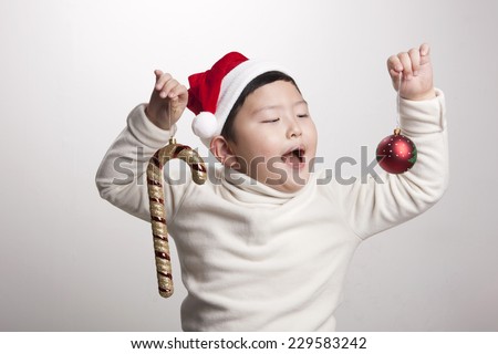 A asian(korea, japan, chinese, china) boy hold red christmas ornaments(decorations) candy, ball and wearing christmas hat with big smile isolated white at the studio.