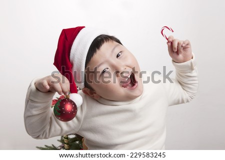 A asian(korea, japan, chinese, china) boy hold red christmas ornaments(decorations) candy, ball and wearing christmas hat with big smile isolated white at the studio.