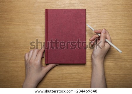 A female(woman) hands hold a blank(empty) red book(note, diary) cover with pencil on the wood desk(table), top view at the studio.