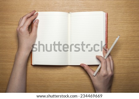 A female(woman) hands hold a blank(empty) red book(note, diary) with pencil on the wood desk(table), top view at the studio.