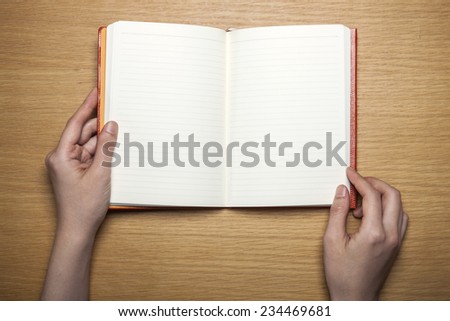 A female(woman) hands hold a blank(empty) red book(note, diary) on the wood desk(table), top view at the studio.