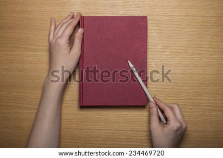 A female(woman) hands hold a blank(empty) red book(note, diary) cover with pencil on the wood desk(table), top view at the studio.