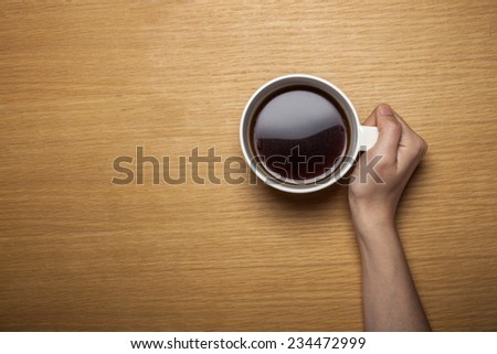 A female hands hold a cup of coffee on the wood table(desk), top view at the studio.