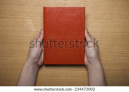 A female(woman) hands hold a blank(empty) red book(note, diary) on the wood desk(table), top view at the studio.
