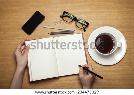 A female hands hold a book and pencil, a coffee and cup and a plate and glasses and smart phone(mobile) on the wood table(desk), top view at the studio.